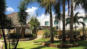 Un jardín cuidado con palmeras, una estructura de techo de paja y un edificio blanco con ventanas.