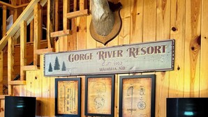 Un mur en bois orné d’une tête de cerf, de dessins de brevets encadrés et d’une enseigne indiquant « Gorge River Resort ».