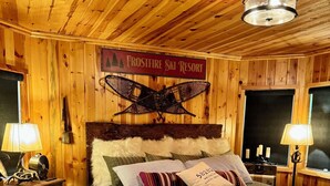 Une pièce chaleureuse aux murs en bois, avec un lit, deux tables de chevet, un banc et une enseigne indiquant « Station de ski Frostfire ».