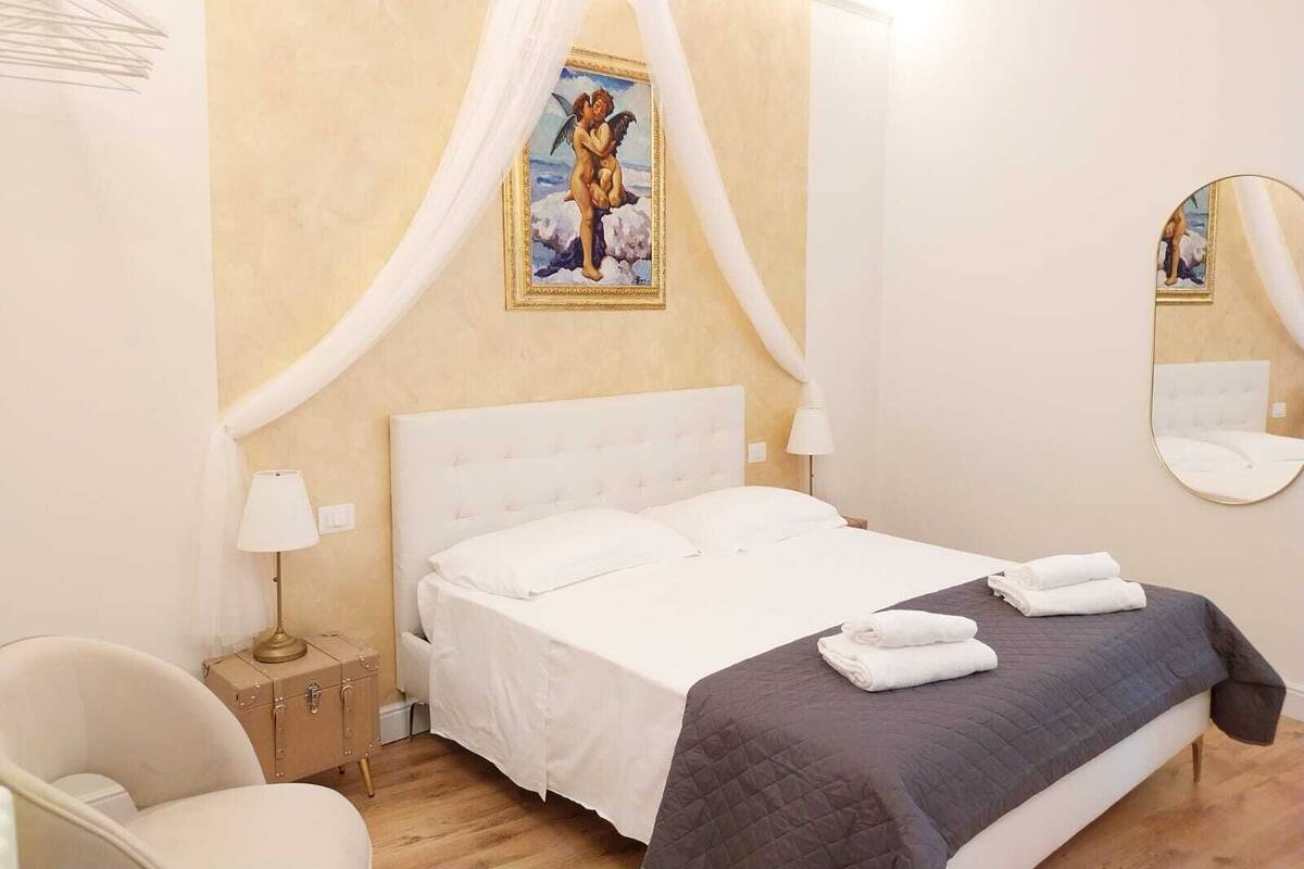 A bedroom with a bed, a bedside lamp, a chair, a nightstand, and a painting on the wall.