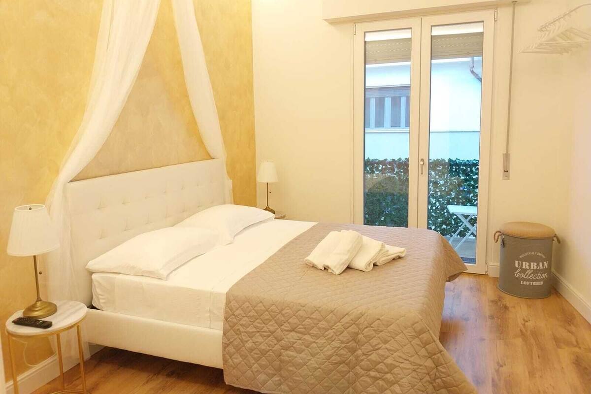 A bedroom with a bed, a bedside table with a lamp, a window with a view, and a trash can.