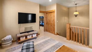 A living room with a flat-screen TV, wooden flooring, a patterned rug, a bench, and a staircase with a wooden railing. - 3-Bed Townhome in Big Sky Town Center (Big Sky)