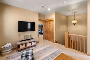 Een woonkamer met een flatscreen televisie, houten vloeren, een kamerkleed met een patroon, een bankje en een trap met een houten leuning.