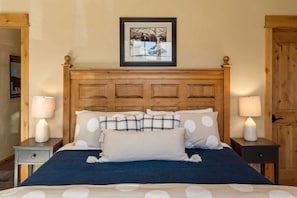 A neatly made bed with a blue bedspread and white pillows, flanked by two bedside tables with lamps, in a room with wooden walls and a framed picture above the bed.