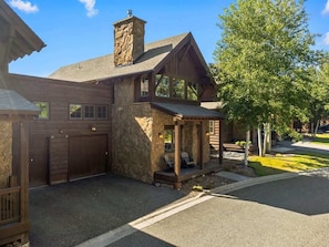 A two-story log cabin with a stone chimney, a covered porch with seating, and a garage.