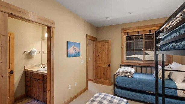 A bedroom with bunk beds, a wooden door, a window, and a bathroom with a sink and mirror. - 3-Bed Townhome in Big Sky Town Center (Big Sky)
