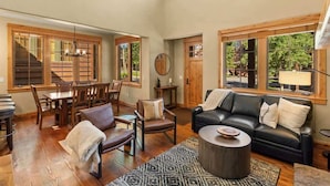 A spacious living room with a black leather sofa, wooden dining table, and chairs, large windows with wooden frames, and a staircase leading to an upper level. - 3-Bed Townhome in Big Sky Town Center (Big Sky)