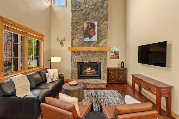 A cozy living room with a stone fireplace, leather furniture, a large window, and a mounted TV.