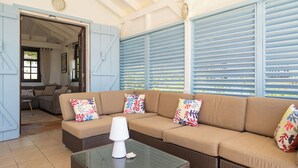 Een woonkamer met een beige hoekbank, gekleurde kussens, een glazen salontafel en blauwe luiken.