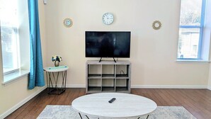 Una sala de estar con un televisor de pantalla plana, una mesa redonda de centro, una mesita auxiliar con un florero y una estantería.