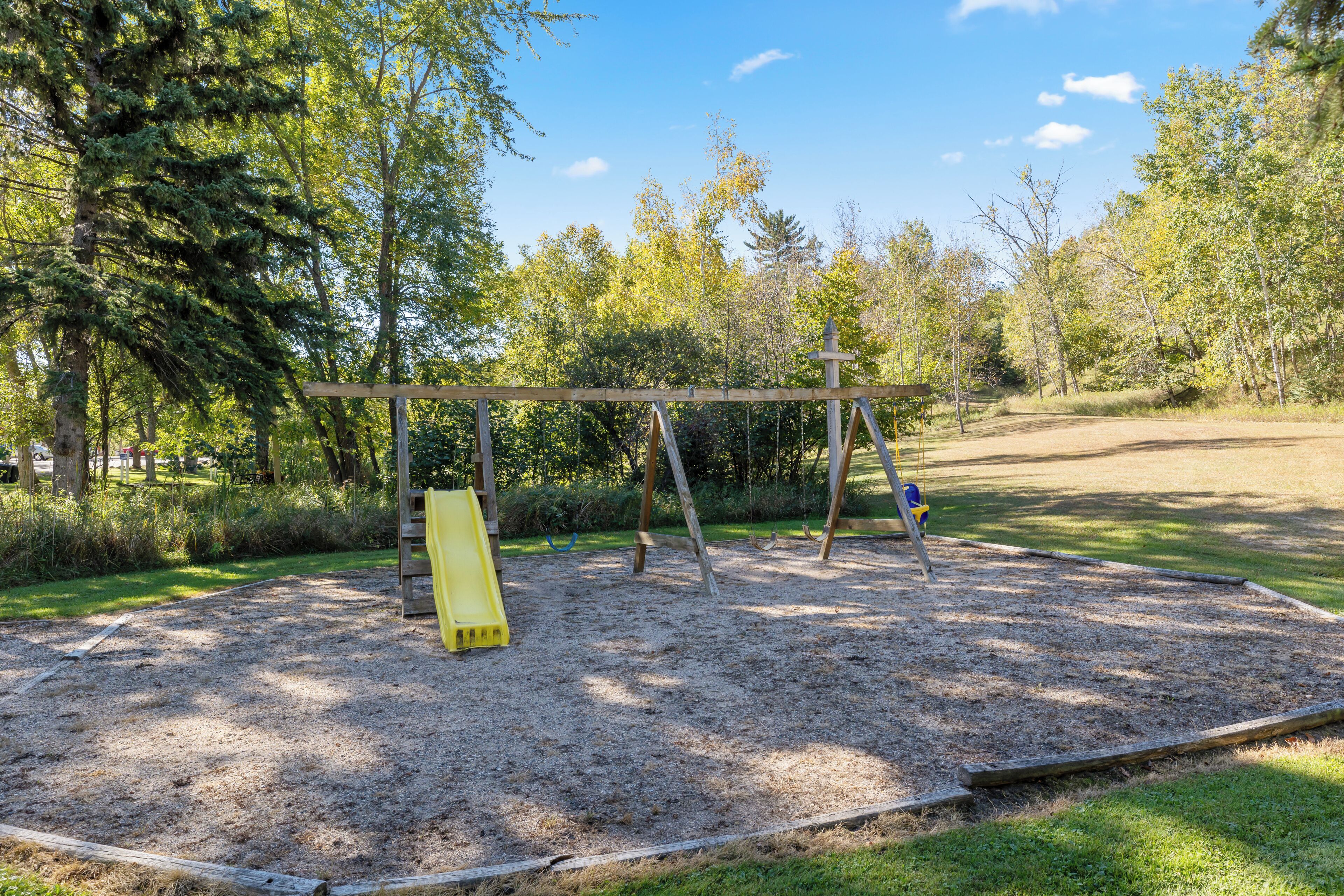 A playground with a swing set and a slide, surrounded by trees and a dirt path.