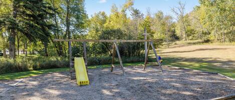 A playground with a swing set and a slide, surrounded by trees and a dirt path.