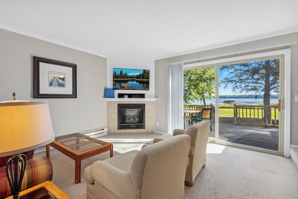 A living room with a fireplace, a sofa, a TV, and a large sliding glass door leading to an outdoor deck with a view of the lake. - North Beach Resort (Park Rapids)