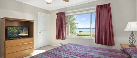 Un dormitorio con una cama grande, un ventilador de techo, un televisor sobre un mueble de madera, una ventana con cortinas y vistas al exterior.