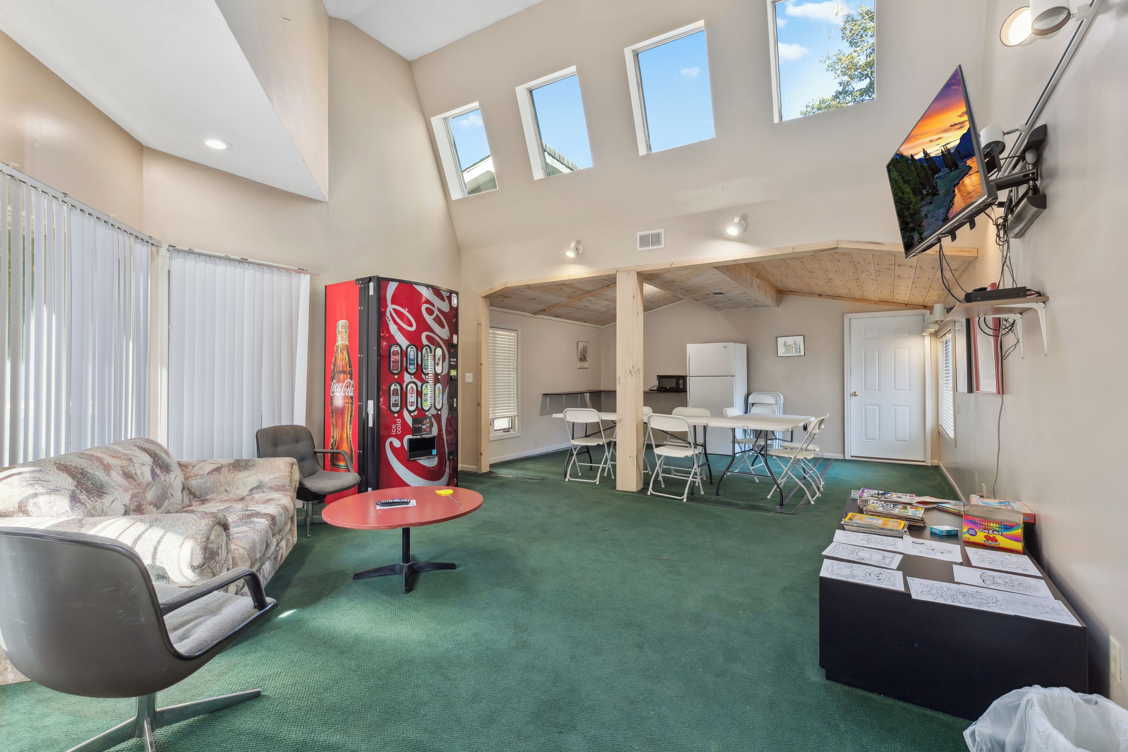 A spacious room with a green carpet, a sofa, a red table, and a Coca-Cola refrigerator. There are windows, a TV, and a desk with chairs.