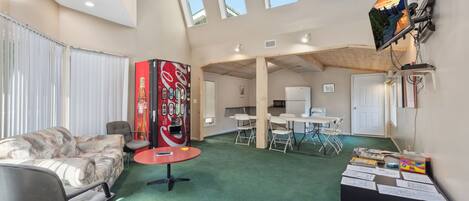 A spacious room with a green carpet, a sofa, a red table, and a Coca-Cola refrigerator. There are windows, a TV, and a desk with chairs.