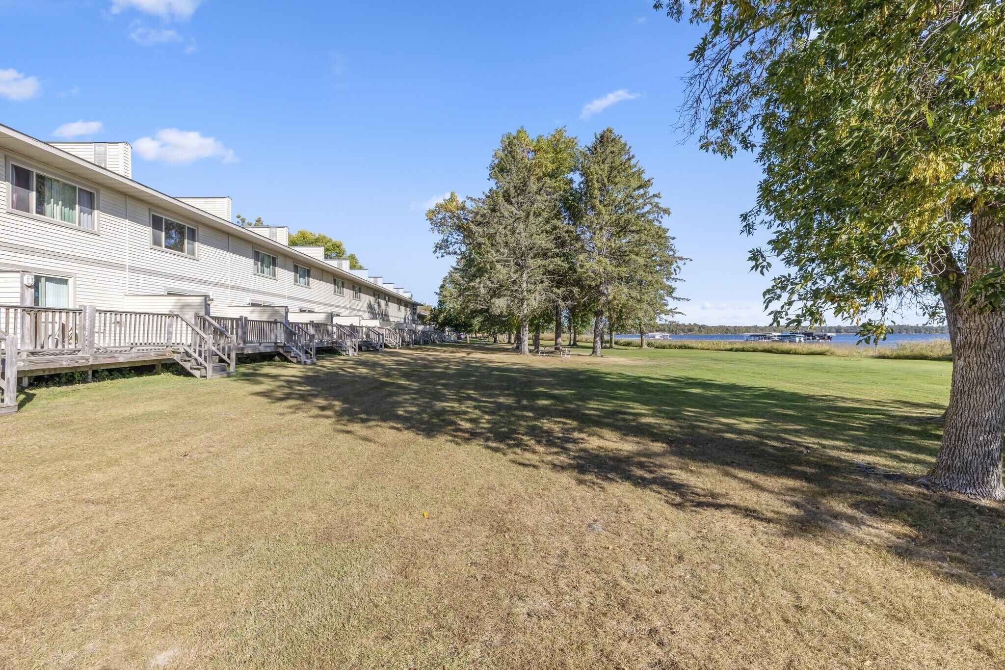 A row of single-story buildings with a grassy lawn and trees in the foreground, and a body of water in the background.