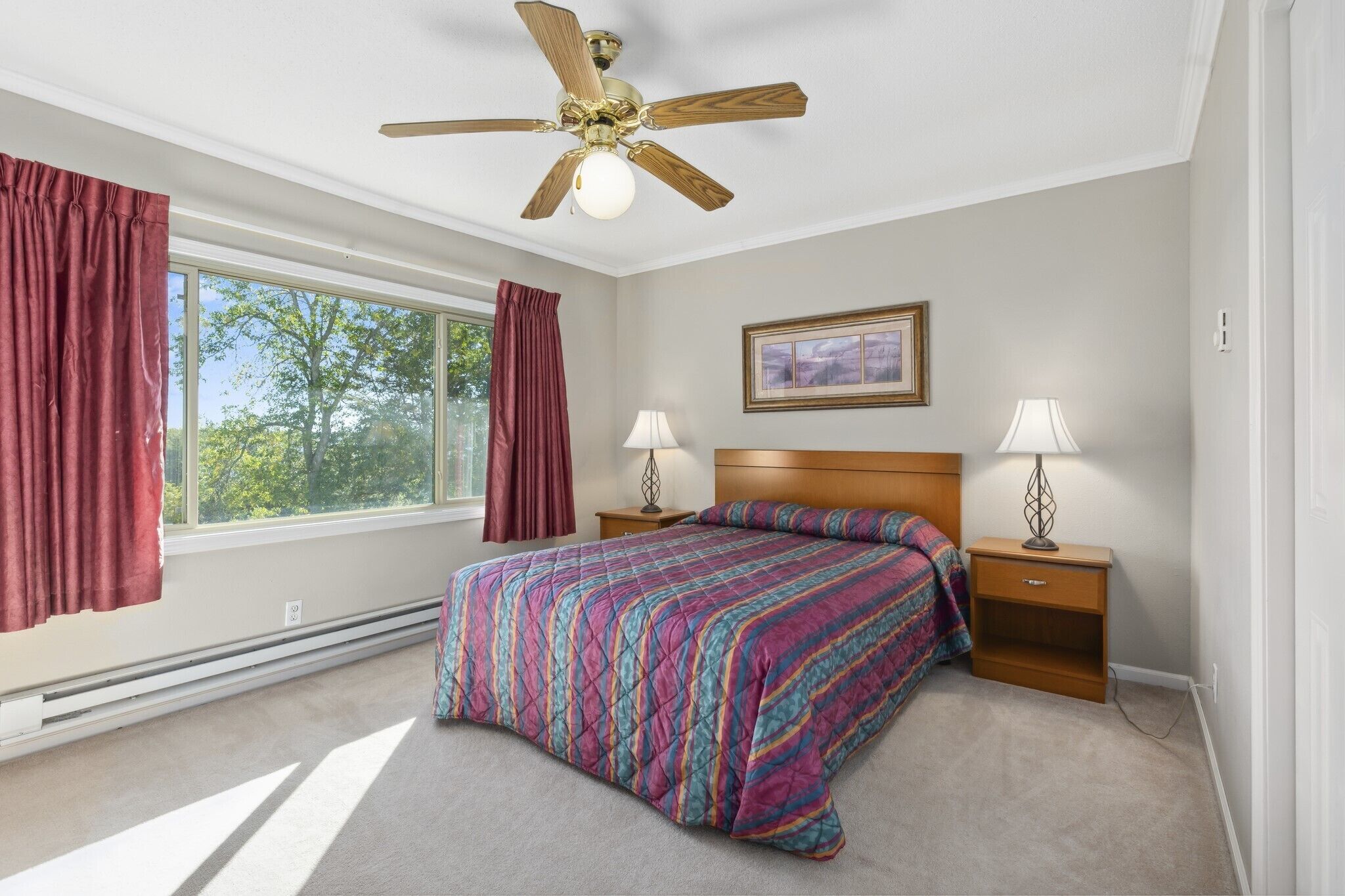 A bedroom with a bed, two bedside tables, a ceiling fan, a window with curtains, and a framed picture on the wall.