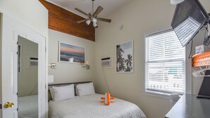 A bedroom with a bed, a ceiling fan, a window with blinds, and a desk with a lamp. - Leo's Lodge | Newly Updated! (Key West)
