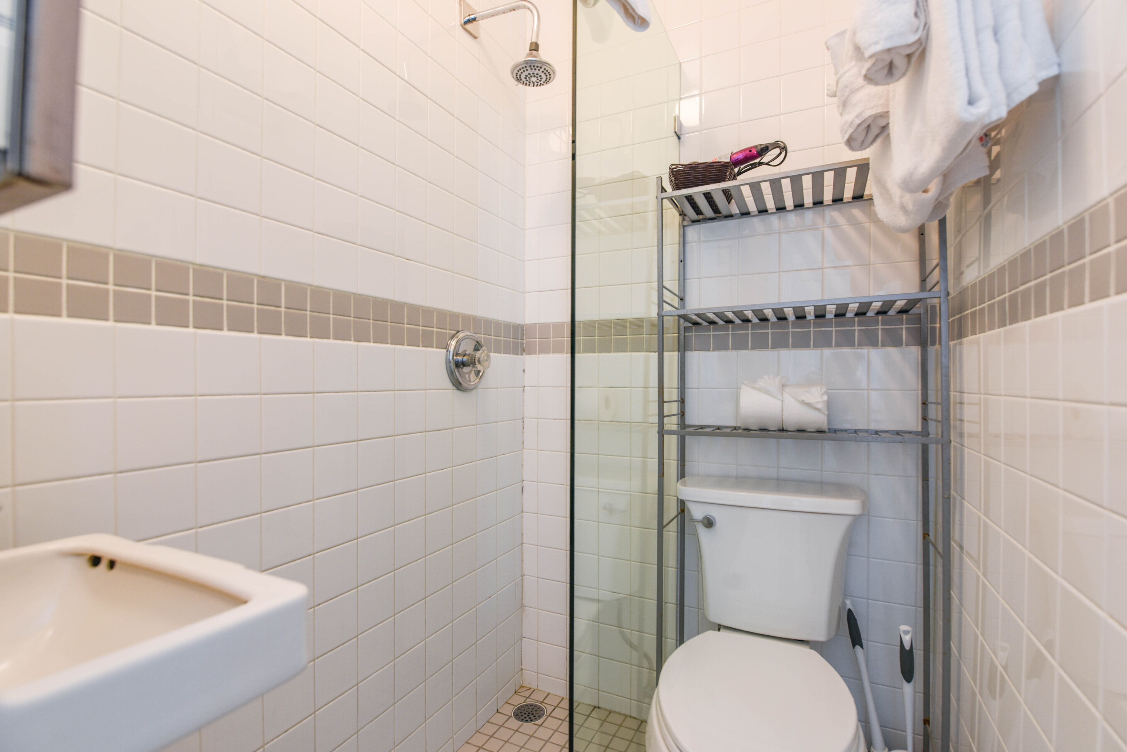 Kylpyhuone, jossa on valkoiset laatat, lasinen suihkuseinä, wc-istuin, pesuallas ja pyyhekoukku.