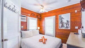 A bedroom with a wooden wall, a bed with white bedding, a ceiling fan, and a framed picture on the wall. - Carey's Corner | Updated Studio with Heated Pool (Key West)