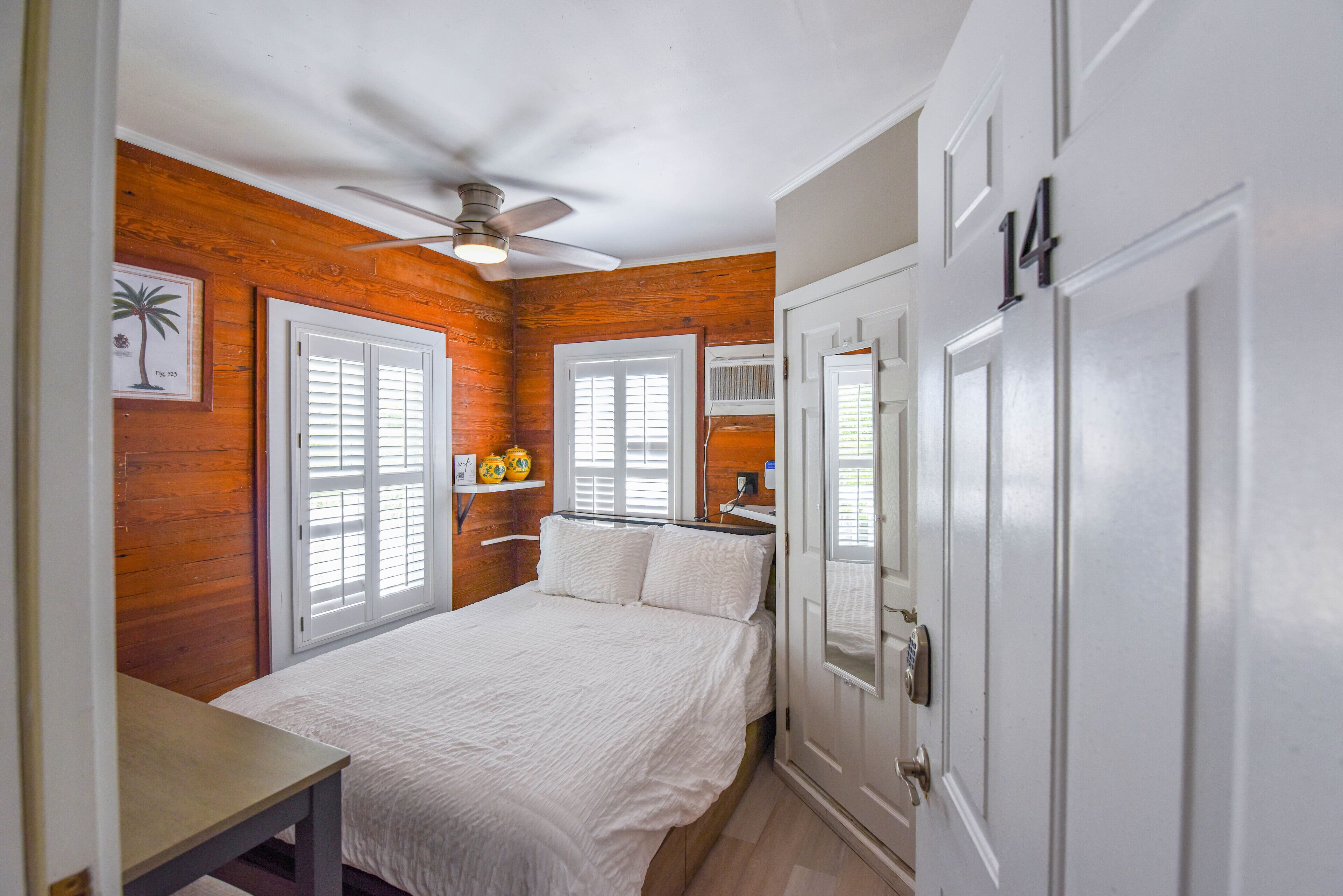 A bedroom with a bed, white walls, wooden paneling, a ceiling fan, and a door with the number 14.