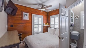 A bedroom with a wooden wall, a bed, a ceiling fan, a bedside table, and a bathroom with a sink and mirror. - Chagall's Corner | Newly Remodeled! (Key West)