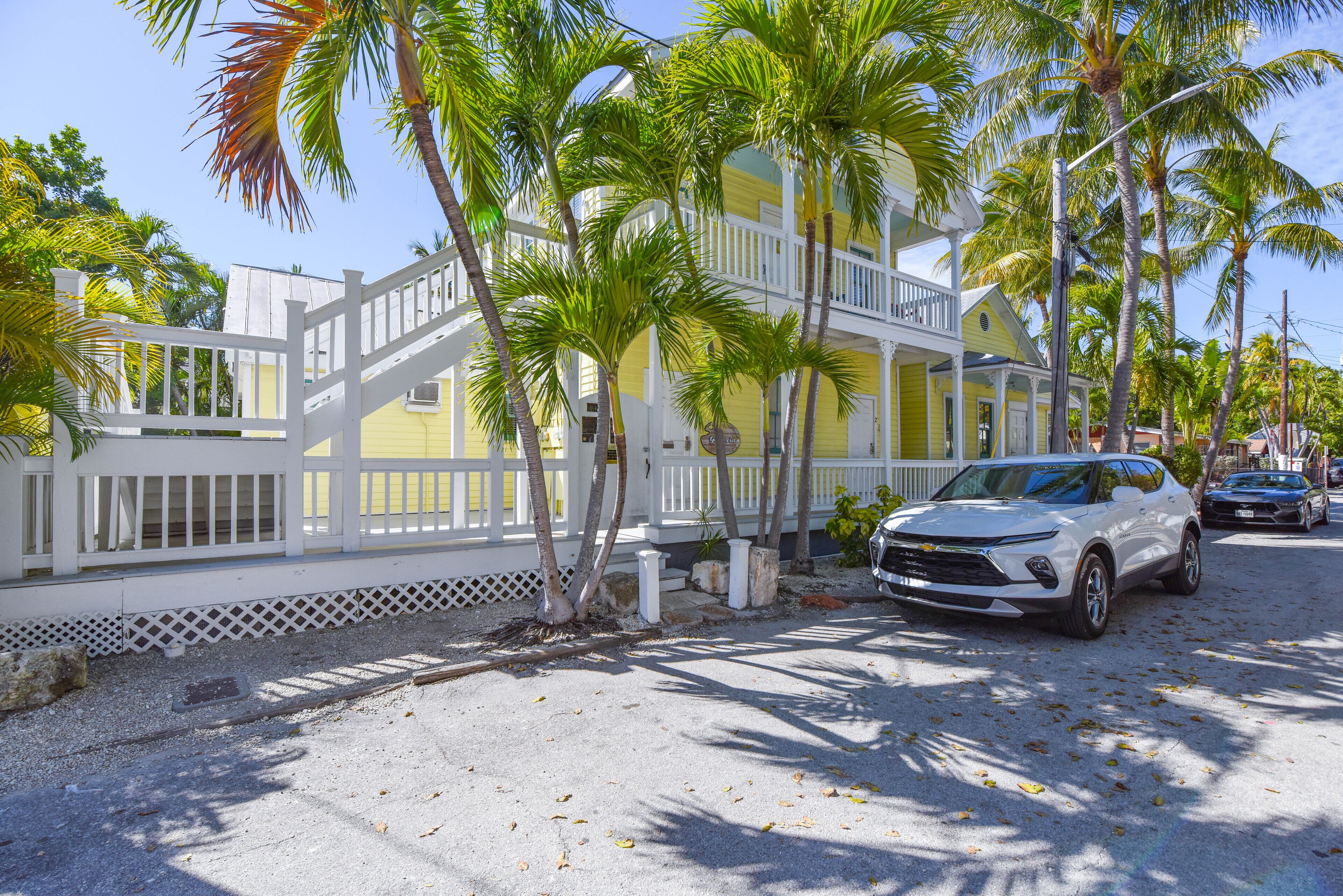 A yellow building with a white balcony, surrounded by palm trees and parked cars.