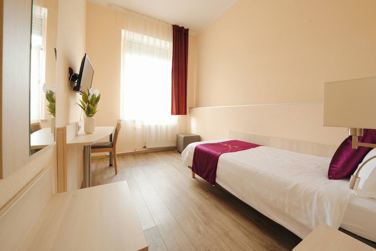 Une chambre d’hôtel avec un lit, un bureau, une chaise, un miroir et une fenêtre avec des rideaux.