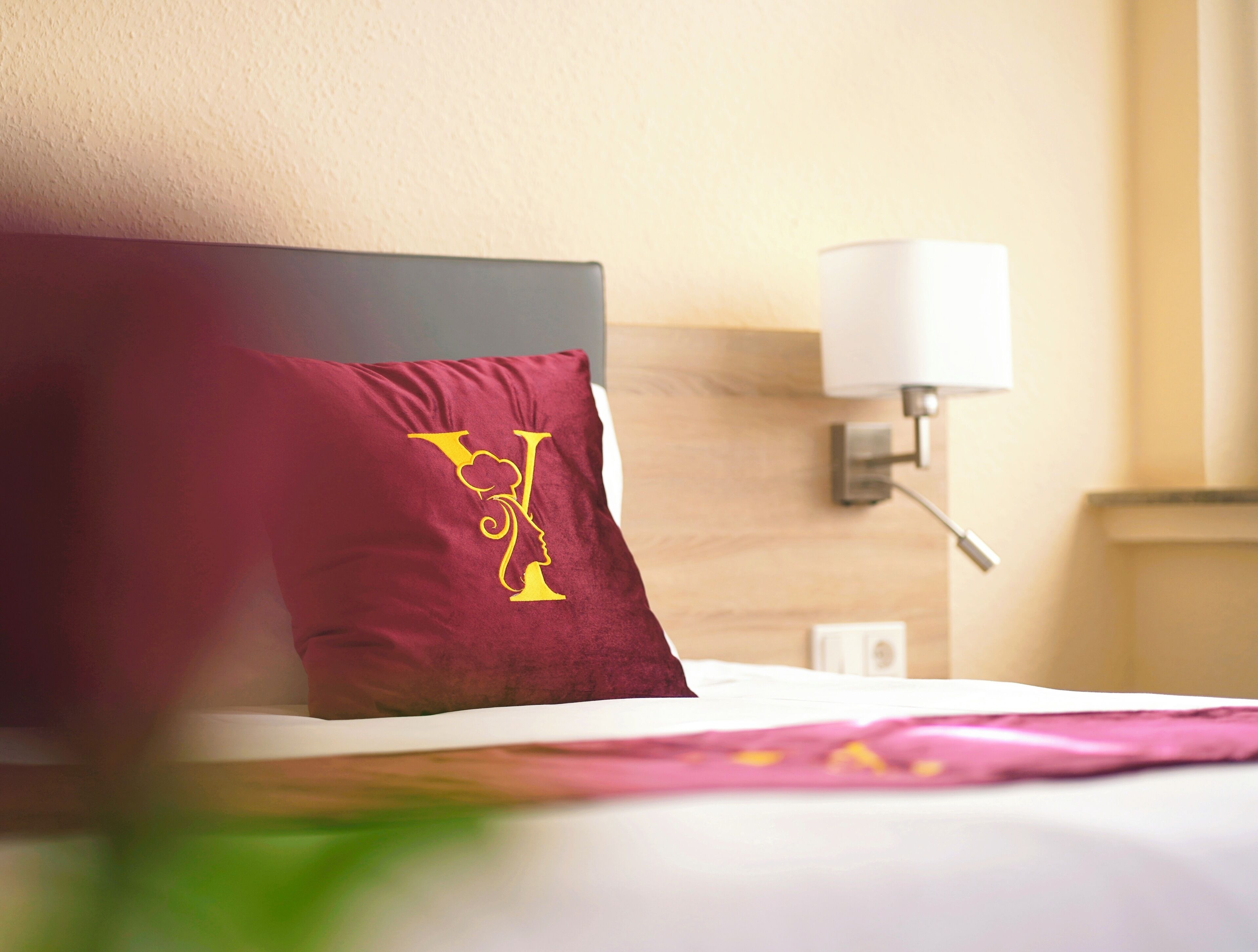 A hotel room with a bed, a red pillow with a gold design, a white lamp, and a wooden headboard.