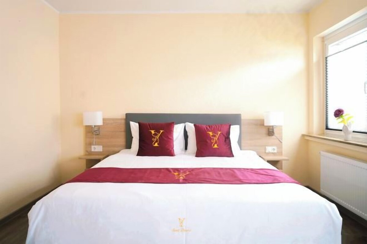A hotel room with a bed, two pillows, a nightstand, a lamp, and a window with a flower.