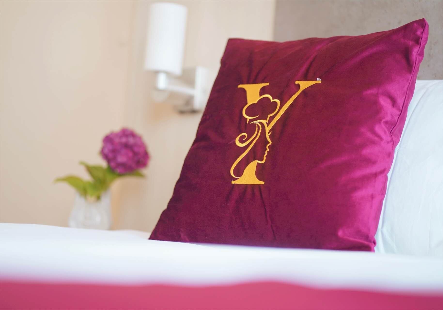 A bed with a white pillow and a maroon pillow with a gold monogram.