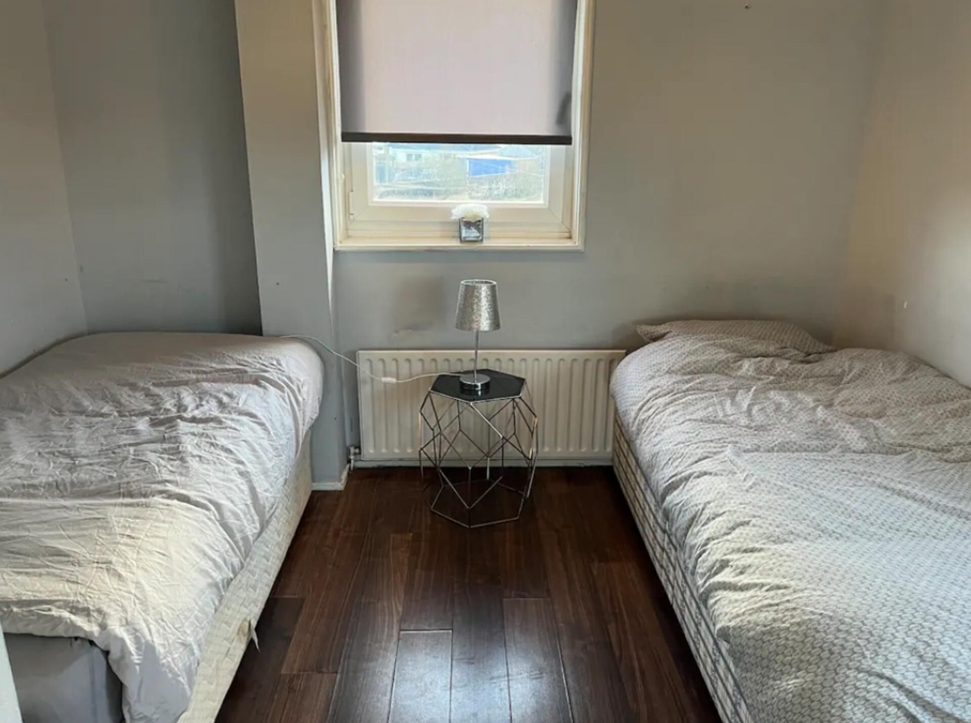 A small, single-room with two beds, a radiator, and a window with a blind.