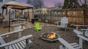 Ein Garten mit überdachter Terrasse, Sitzgelegenheiten im Freien, einer Feuerstelle und einem Holzzaun.