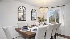 Ein Esszimmer mit einem rechteckigen Tisch, grauen Polstersesseln, einer Kristallleuchter und einem großen Fenster mit Vorhängen.