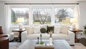 Ein Wohnzimmer mit einem weißen Sofa, zwei Beistelltischen mit Lampen, einem Couchtisch mit Dekoration und Blick auf Bäume und Häuser durch große Fenster.