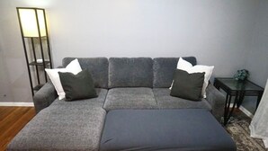 A grey sectional sofa with a matching ottoman, two white and two dark grey cushions, a floor lamp, and a small table with a vase. - Peaceful Home In The Heart of TLH (Tallahassee)