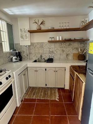 Eine Küche mit weißen Schränken, einem Herd, einer Spüle und einem Kühlschrank. Ein Fenster mit Jalousien, eine Kaffeemaschine und ein Teppichboden sind ebenfalls vorhanden.