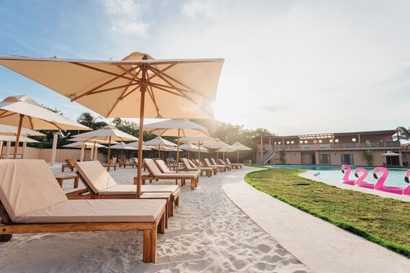 A poolside area with sun loungers and umbrellas, a pool with pink flamingo floats, and a building in the background.