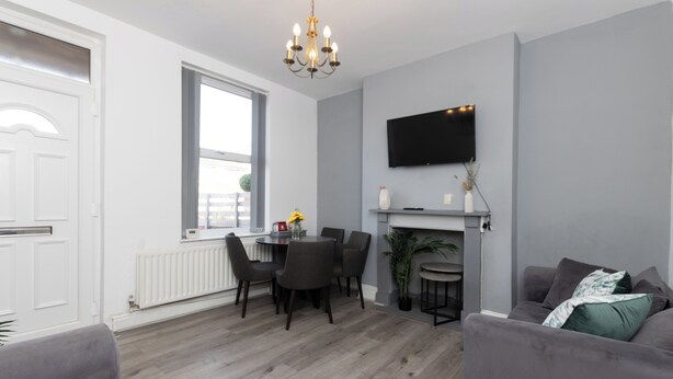 A modern living room with a grey sofa, a dining table with chairs, a chandelier, a fireplace, and a TV mounted on the wall.