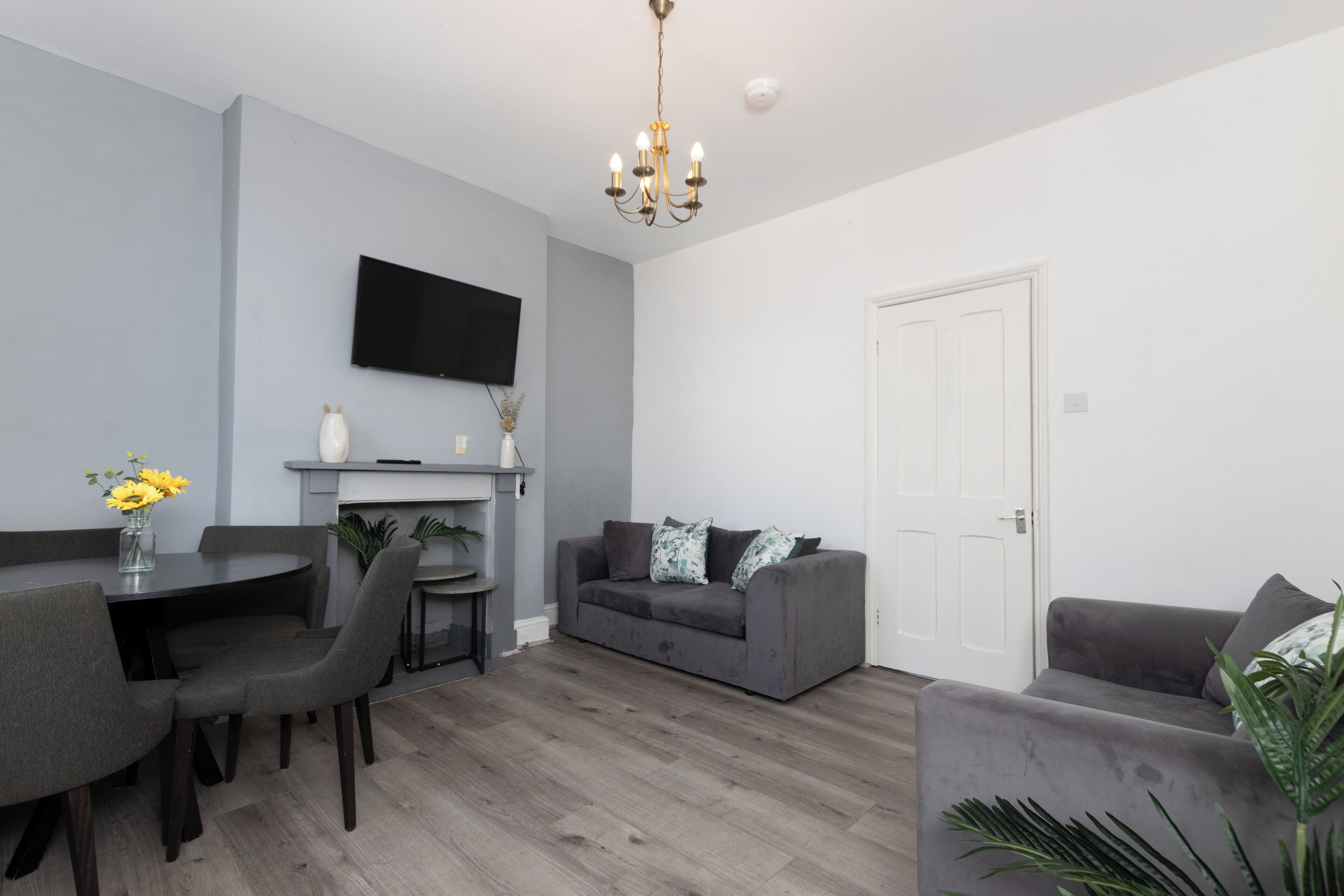Un salon moderne avec un canapé gris, une table à manger avec des chaises, un téléviseur à écran plat fixé au mur et un lustre.