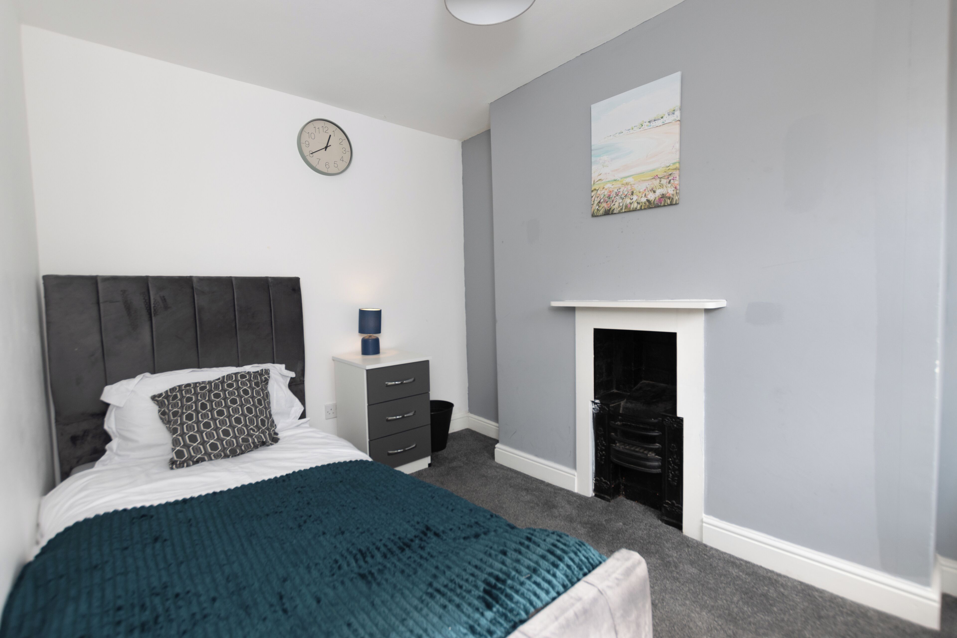 A bedroom with a bed, a dresser, a clock, a fireplace, and a painting on the wall.