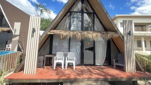 A modern, triangular-roofed building with a thatched roof entrance, a wooden deck, and white plastic chairs.