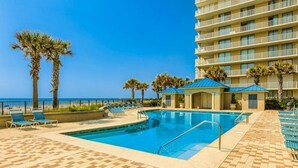 A hotel pool area with a clear view of the ocean, palm trees, and a building with balconies. - Bluewater 1501 | Beach Front (Orange Beach)