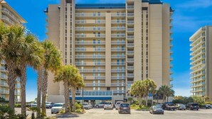 A multi-story hotel with a central atrium, palm trees, and a parking lot. - Bluewater 1501 | Beach Front (Orange Beach)