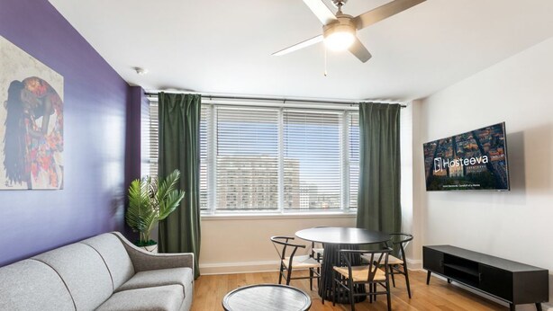 A living room with a sofa, a round table, a ceiling fan, a TV, and a window with blinds.