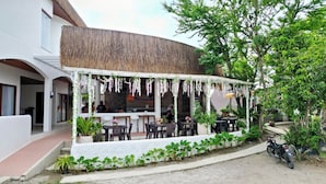 An outdoor seating area with tables and chairs, surrounded by potted plants and a thatched roof structure.