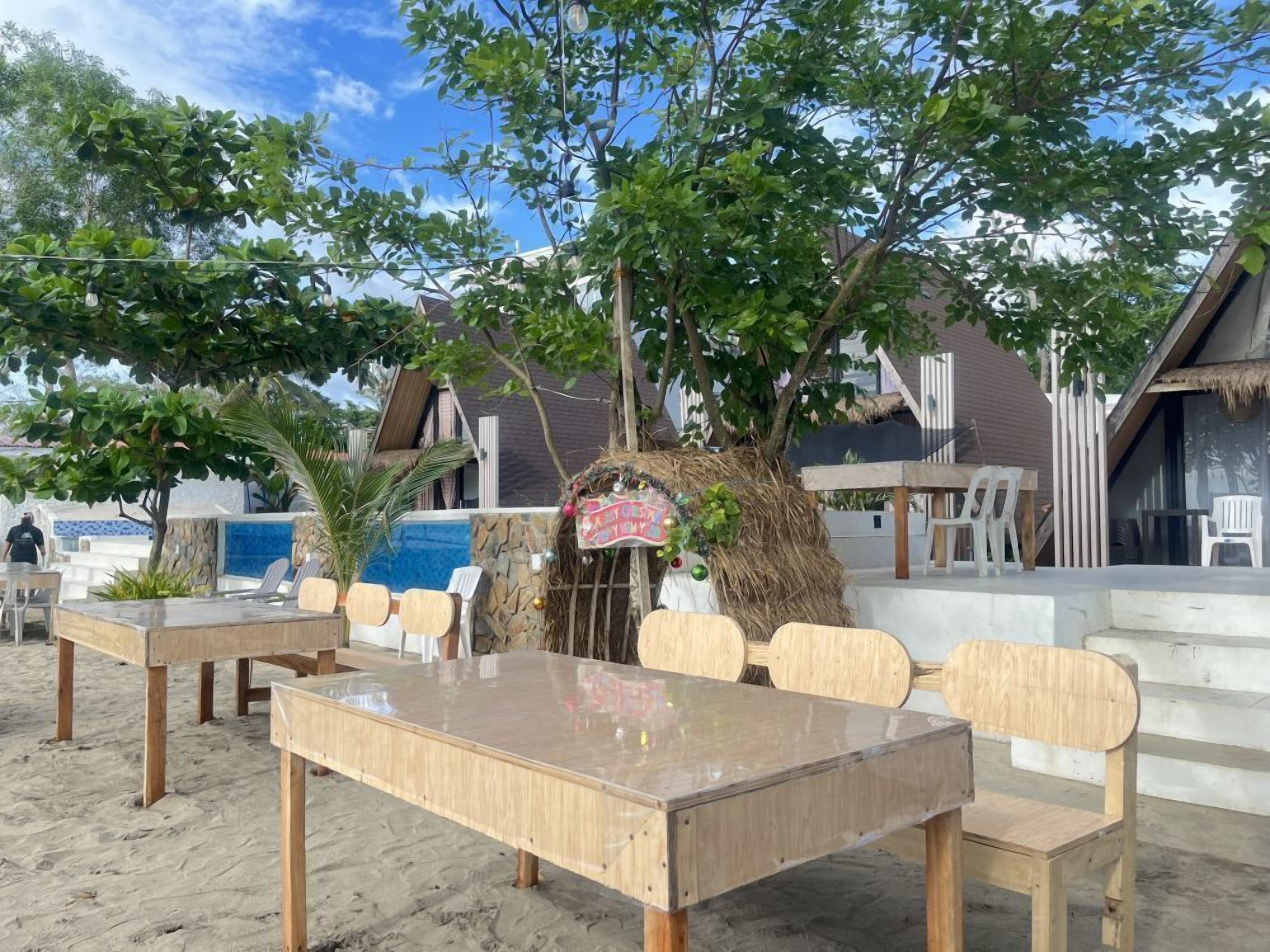 Un espace de restauration situé en bord de mer, avec des tables et des chaises en bois, une structure dotée d’un toit de chaume et un panneau indiquant la présence d’un restaurant.