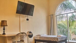 A room with a bed, a desk, a chair, a TV, and a large window with a view of palm trees. - Jardin Zen  (Tepoztlán)