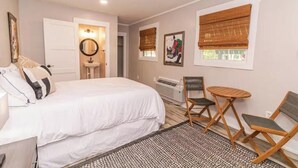 Une chambre à coucher comprenant un lit, une table de chevet, une chaise, une petite table et une fenêtre avec un store.